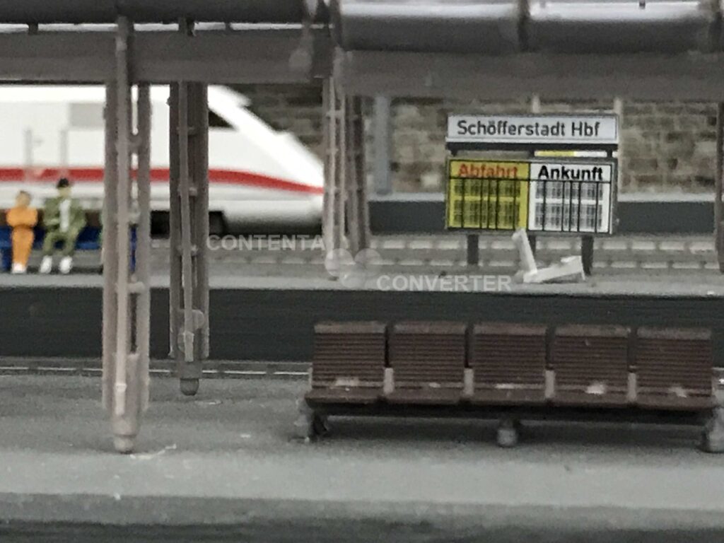 Auf dem hinteren Bahnsteig ist der Triebwagen eines ICEs zu sehen.
Im Vordergrund steht eine Bank. Links im Bild sitzen zwei Reisende auf einer Bank und warten.