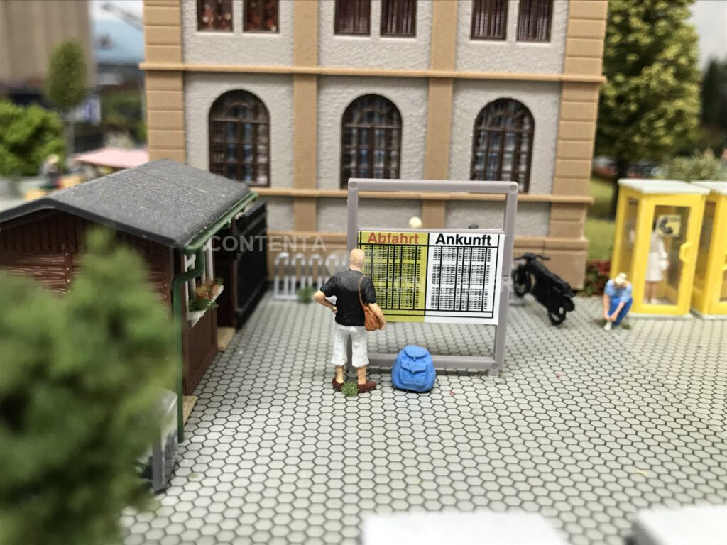 Ein Reisender steht vor der Ankunft-/Abfahrtstafel und studiert die Zeiten. Im Hintergrund bindet sich eine Frau ihren Schuh. Eine andere steht in einer Telefonzelle und telefoniert.