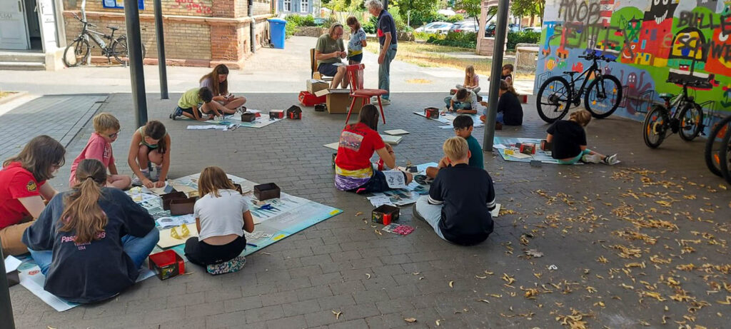 Kinder sitzen im Schulhof beim Basteln der Modellhäuser